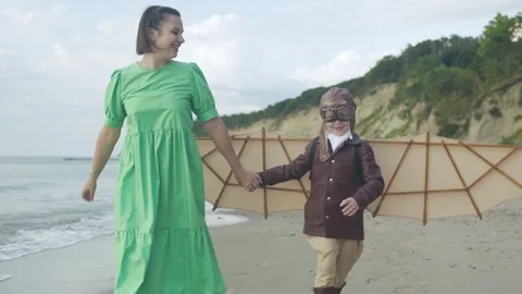 Little boy in pilot suit with artificial wings walks with his mother on seashore Stock Footage 279898235