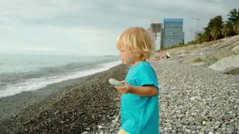 A little boy is playing on the seashore, he throws stones into the water. Family Stock-Footage 200807386