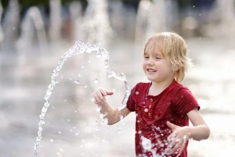 Little boy plays in the square between the water jets in the fountain at sunn Stock Photos