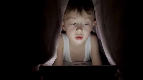 Little boy plays on tablet while lying under blanket. watching cartoons hiding Stock Footage 125787641