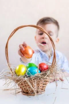 Little boy puts an orange Easter egg in a basket from the table on a white ba 스톡 사진