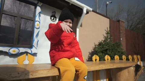 A little boy in a red jacket and yellow pants sits on a bench in the park  Stock Footage 271319554