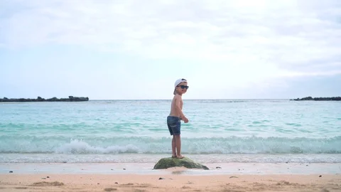 A little boy in shorts, a baseball cap and sunglasses is standing on a stone in Stock Footage 101757523