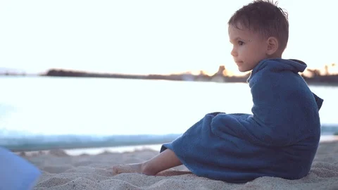 A little boy sits on a sandy seashore wrapped in a towel and basking after Stock Footage 101759768