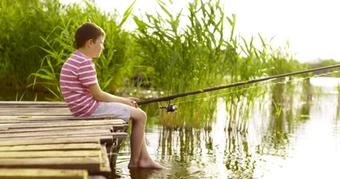 Little boy sitting on the edge of a wooden dock and fishing in lake at sunset Stock Footage