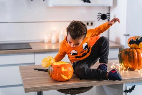 Little boy sitting at kitchen table dressed as pumpkin playing with decorated Stock Photos