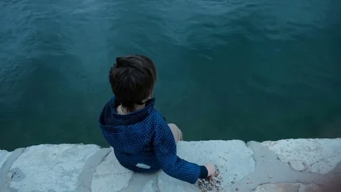 Little boy sitting on pier throws stones into water outdoors Stock Footage 75739276