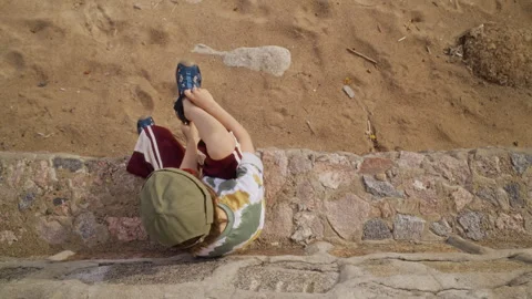 Little Boy Sitting on Stone Ledge Adjusting Sandals above Beach Stock Footage 340256130