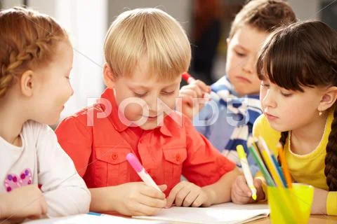 A little boy sitting at table and drawing and his friends looking at him Stock Photos