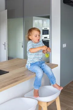 Little boy is sitting on a table in the kitchen and eating grapes. Stock Photos