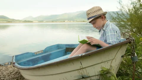 Little boy in a straw hat sitting in the rustic boat on the lake bank and readin Video stock 248929112