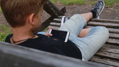 Little boy using smart phone and sitting on the bench in park Stockbeeldmateriaal 115078381