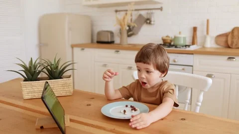 A little caucasian boy eats krunchy and watches tablet PC at kitchen table. Vidéo 171187704