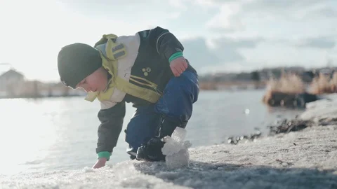 A little Caucasian boy in a hat, jacket and boots plays on the winter bank of Stock Footage 128236166