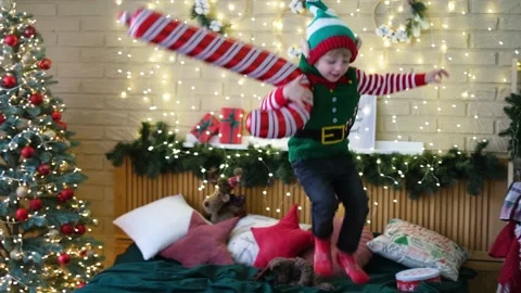 Little cheerful boy in an elf hat jumping on the bed with an inflatable cane Stock Footage 165389996