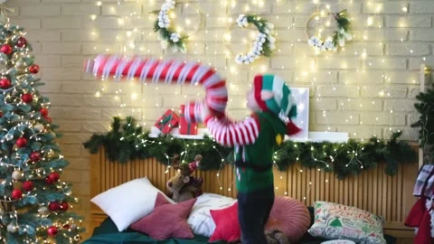 Little cheerful boy in an elf hat jumping on the bed with an inflatable cane Stock Footage 165866698