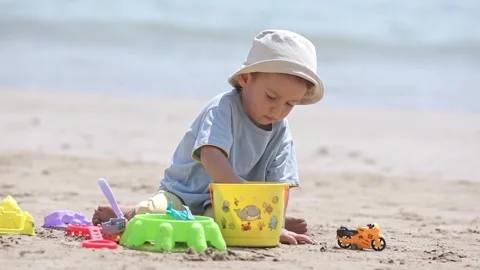 Little child boy sits on sandy beach, plays with bucket and colorful sand toys Stock Footage 332030928