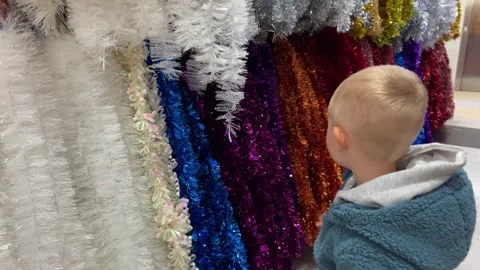 Little curious boy looking and touching multicoloured Christmas tinsel in a shop Stock Footage 322028455