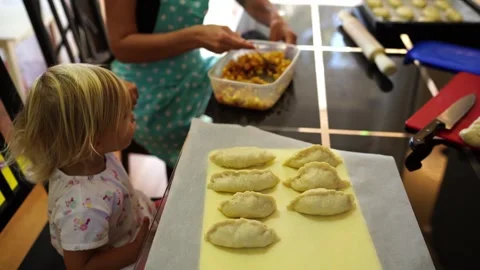 Little daughter eats stuffing with a spoon next to mom filling dough for buns Stock Footage 280343310