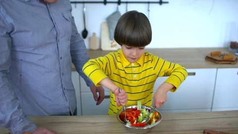 Little funny boy sitting in kitchen and playing with vegetable salad just Stock Footage 83430481