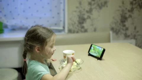 A little girl eats mashed potatoes and watches cartoons on her smartphone Stock Footage 143897057