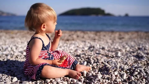 Little girl eats a pancake while sitting on a pebble beach and looking at the Stock Footage 285541471