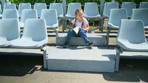 Little girl elementary student with a backpack sitting on a bench and reading a  Stock Footage 280209677