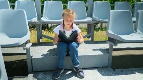 Little girl elementary student with a backpack sitting on a bench and reading a  Stock Footage 281103530