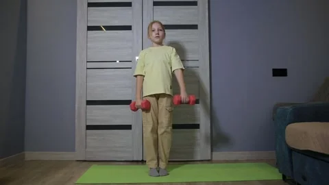 Little girl performs front shoulder raises with dumbbells on a yoga mat at home, Stock Footage 339865732