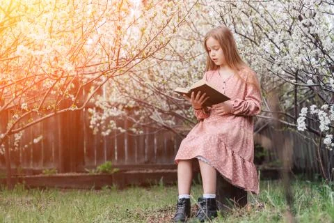 Little girl is reading a book near a flowering tree in the garden. Stock Photos