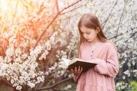 Little girl is reading a book near a flowering tree in the garden. Stock Photos