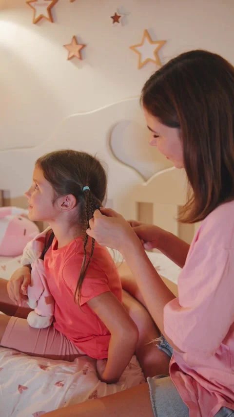 Little girl sits on bed while her mother styles hair in a cozy bedroom during Stock Footage 330746363