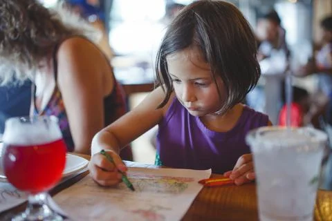 A little girl sits at a restaurant table coloring with crayons Stock Photos