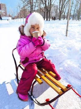A little girl sits on a sled and drinks hot cocoa. A beautiful winter day Stock Photos