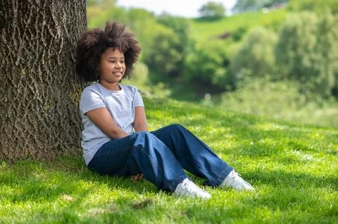 Little girl sitting on grass under tree resting in nature enjoying freshair. Stock Photos