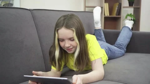 A little girl is smiling while lying on the couch with a tablet. 動画素材 169994697