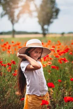 Little longhaired girl in hat posing at field of poppies with  on summer sun. Stock Photos