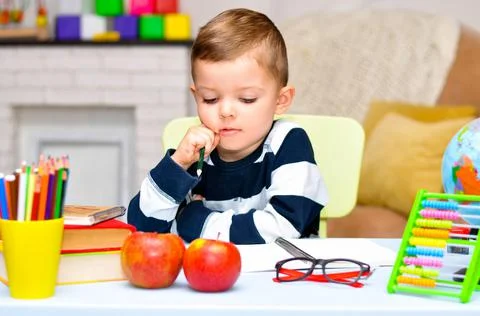 A little preschooler boy is bored and he sits at a table with a book, a globe, a Stock Photos