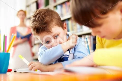 A little schoolboy sitting at table and looking at his working classmate Stock Photos