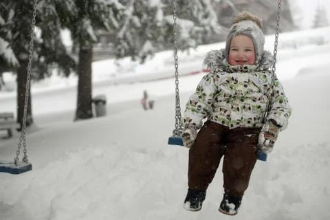 Little smiling child with red cheeks sitting on swing under snowfall, background Stock Photos