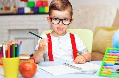 A little surprised preschooler sits at a desk in a white T-shirt and red suspend Stock Photos