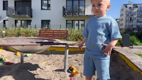 Little two-year-old boy looks around confusedly while standing in a sandbox Stock Footage 307128357