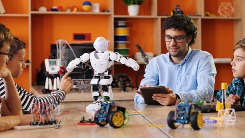 Little white robot walking in front of the kids and their male teacher on the Stock Footage 82481898