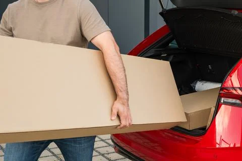 Loading large cardboard box into red hatchback car at outdoor parking area Foto stock