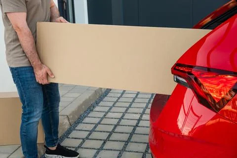 Loading large cardboard box into red car trunk in residential driveway 스톡 사진