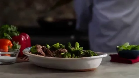 Lock down shot of beef with broccoli with chef flambeing wok in the background Stock Footage 98247769