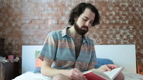 Lockdown shot of Caucasian young man reading book while sitting on bed Stock Footage 114859504