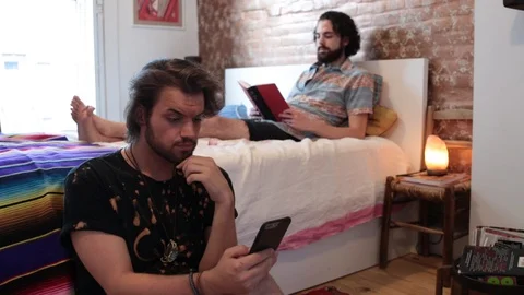 Lockdown shot of gay man using smart phone while boyfriend reading book Stock Footage 114859428