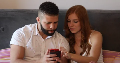 Lockdown shot of young couple using smartphone together while sitting on bed Stock Footage 119977668