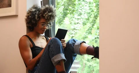 Lockdown shot of young woman using digital tablet while sitting on window sill Vídeos de archivo 119790375